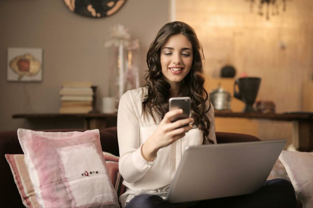 Frau sitzt mit Telefon und Laptop auf einem Sofa.